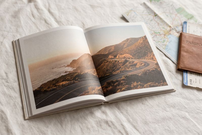 Open travel photo book showing a coastal road at sunset, beside a passport and boarding pass